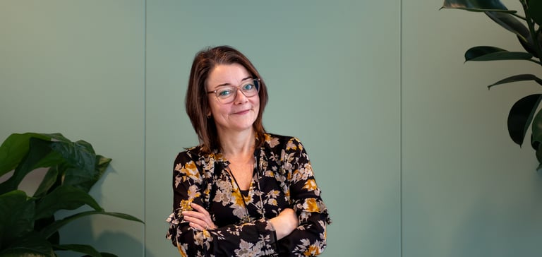 En selvsikker kvinne står med armene i kors foran en lysegrønn vegg, omgitt av planter