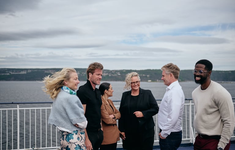 Gruppe snakker på båtdekk med hav og åser i bakgrunnen